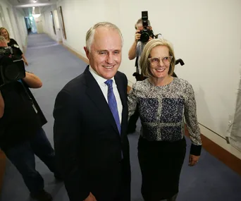 A man in a suit and a woman in glasses smile as they walk down a hallway, followed by photographers.
