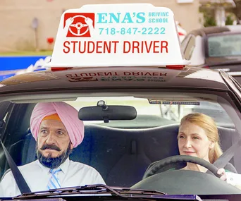 A woman in a driver's seat and a man with a turban sit in a car with a "Student Driver" sign on top.