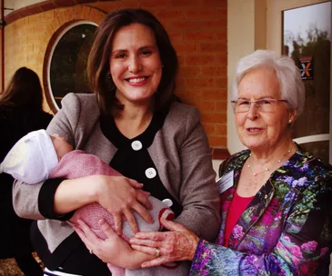 A woman holds a baby, standing beside an elderly woman, both smiling outdoors.