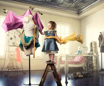 A young girl dressed as a fashion designer stands on a ladder, styling a mannequin with fabric in a bright studio.