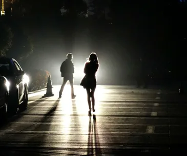 Silhouettes of two people walking on a dark street, illuminated by headlights, casting long shadows.