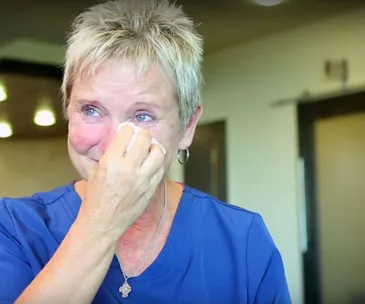 A woman in a blue shirt wipes tears from her eyes, appearing emotional in a well-lit room.