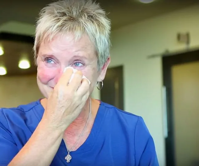 A woman in a blue shirt wipes tears from her eyes, appearing emotional in a well-lit room.