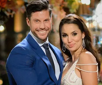 A man in a blue suit and a woman in a white dress, both smiling, posing together with a blurred floral backdrop.