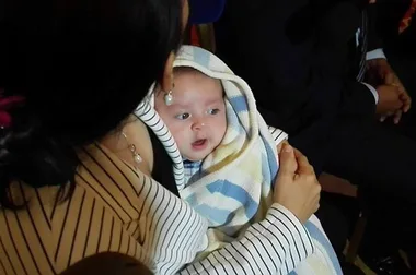 A baby wrapped in a blanket is held by a woman in a striped outfit, with other people visible in the background.