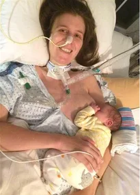Mother in hospital bed breastfeeding her baby, with medical tubes attached, smiling despite her condition.