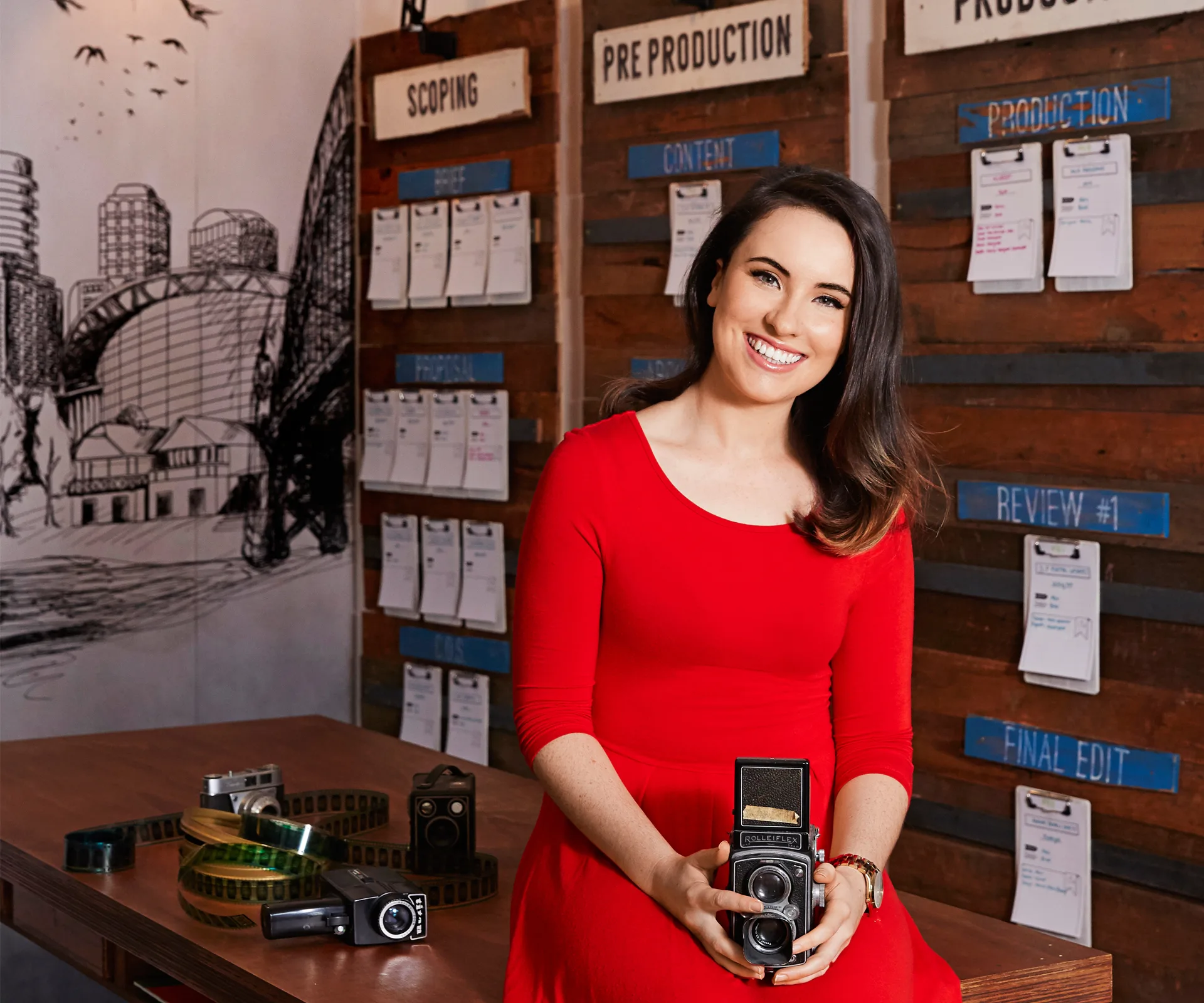 Woman in a red dress holding a vintage camera, sitting in a creative studio with storyboards and film equipment.