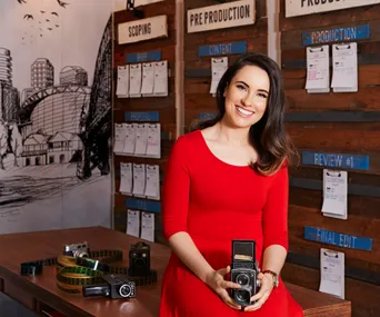 Woman in a red dress holding a vintage camera, sitting in a creative studio with storyboards and film equipment.
