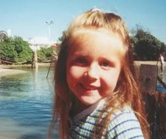 A young girl with long hair, smiling outdoors near a body of water and greenery.