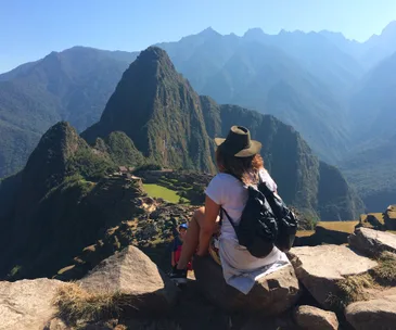 A person with a hat and backpack sits on a rock, overlooking Machu Picchu with mountains in the background.