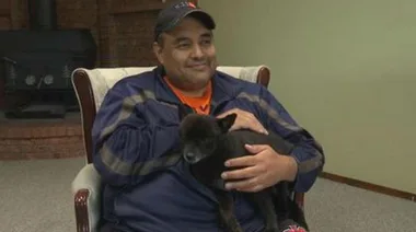 Man sitting on a chair, smiling and holding a small black dog in his arms, indoors.