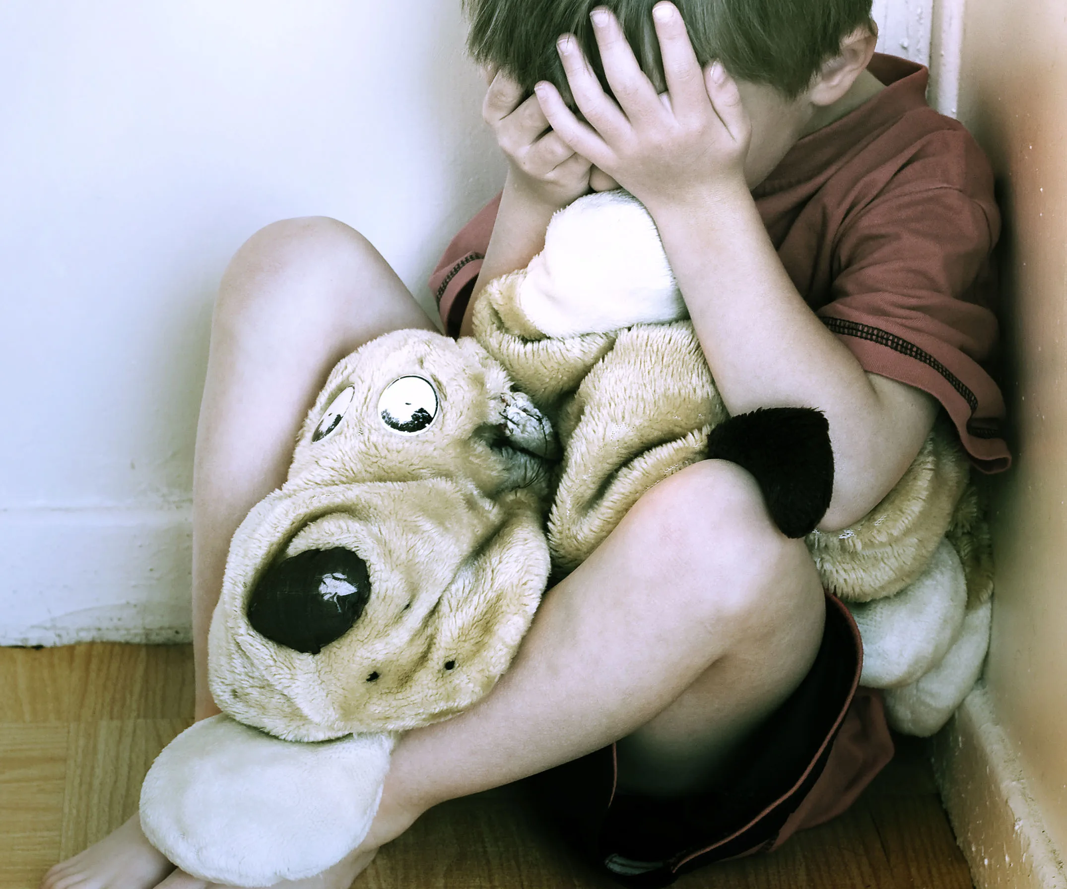 A child sits in a corner, holding a large stuffed animal and covering their face with their hands.
