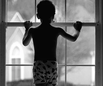 A silhouetted child in shorts stands at a window, gazing outside.