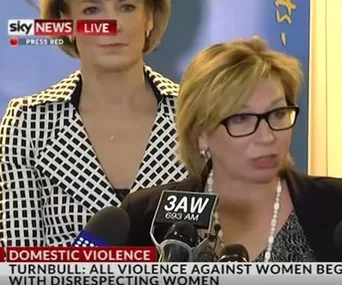 Rosie Batty speaking at a press conference on domestic violence, with microphones in front, another woman stands behind.