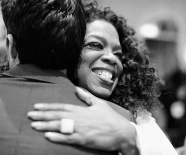 A woman with curly hair smiling widely while hugging another person.