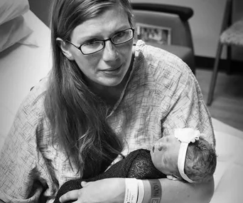 A mother in a hospital gown holds her newborn baby, wrapped in a blanket, with a gentle expression.