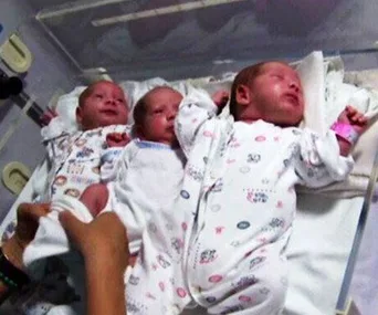 Three newborn triplets in hospital bassinets wearing white sleepers with colorful patterns.