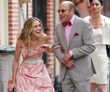 Smiling woman in a pink floral dress with a man in a gray plaid suit and bow tie, walking arm-in-arm on a city street.