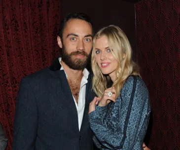 A couple posing together, man with beard in a suit, and woman with blonde hair, in front of a patterned red wall.