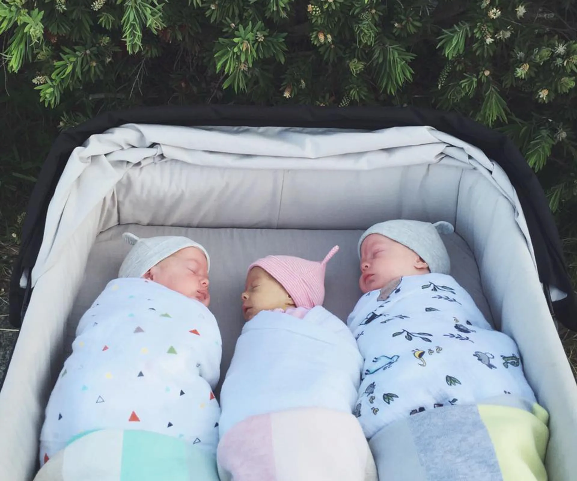 Three newborn babies, swaddled in patterned blankets and tiny hats, sleep peacefully in a stroller outdoors.