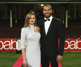 A couple poses elegantly on a red carpet at an outdoor event, with stadium seating in the background.