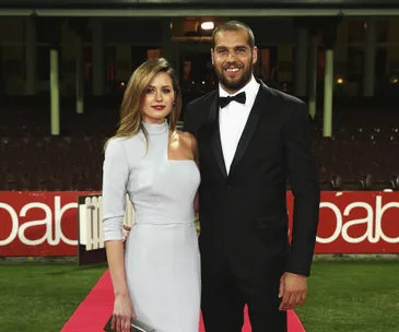 A couple poses elegantly on a red carpet at an outdoor event, with stadium seating in the background.