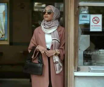 A woman in a hijab and sunglasses stands outside a building, wearing a brown coat and holding a black handbag.