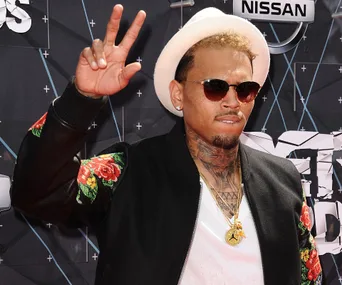 A man wearing sunglasses and a hat, making a peace sign on a red carpet.