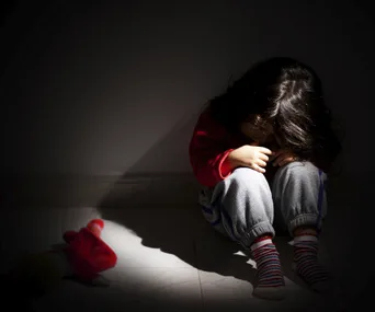 Child sitting alone in shadow on floor, head down, with a toy nearby, conveying distress.