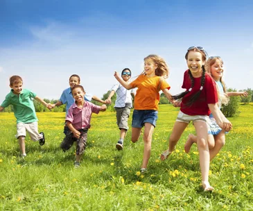 Children running joyfully in a sunny field, holding hands, surrounded by green grass and yellow flowers.