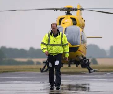 Man in yellow jacket walking on wet tarmac in front of a yellow helicopter in a cloudy setting.
