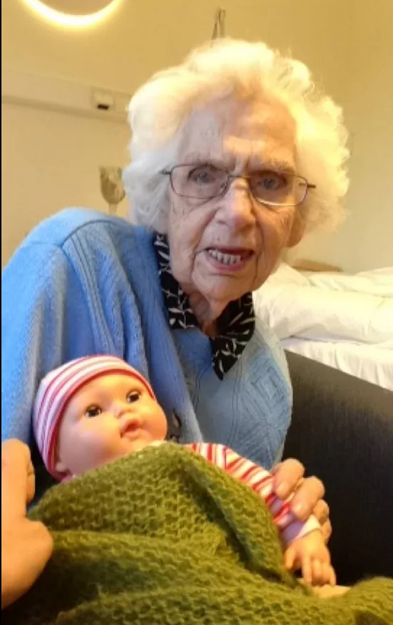 Elderly woman with glasses holding a doll wrapped in a green blanket, wearing a blue sweater in a cozy room.
