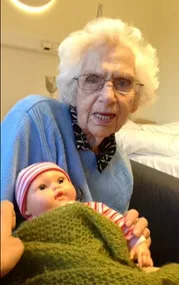 Elderly woman with glasses holding a doll wrapped in a green blanket, wearing a blue sweater in a cozy room.