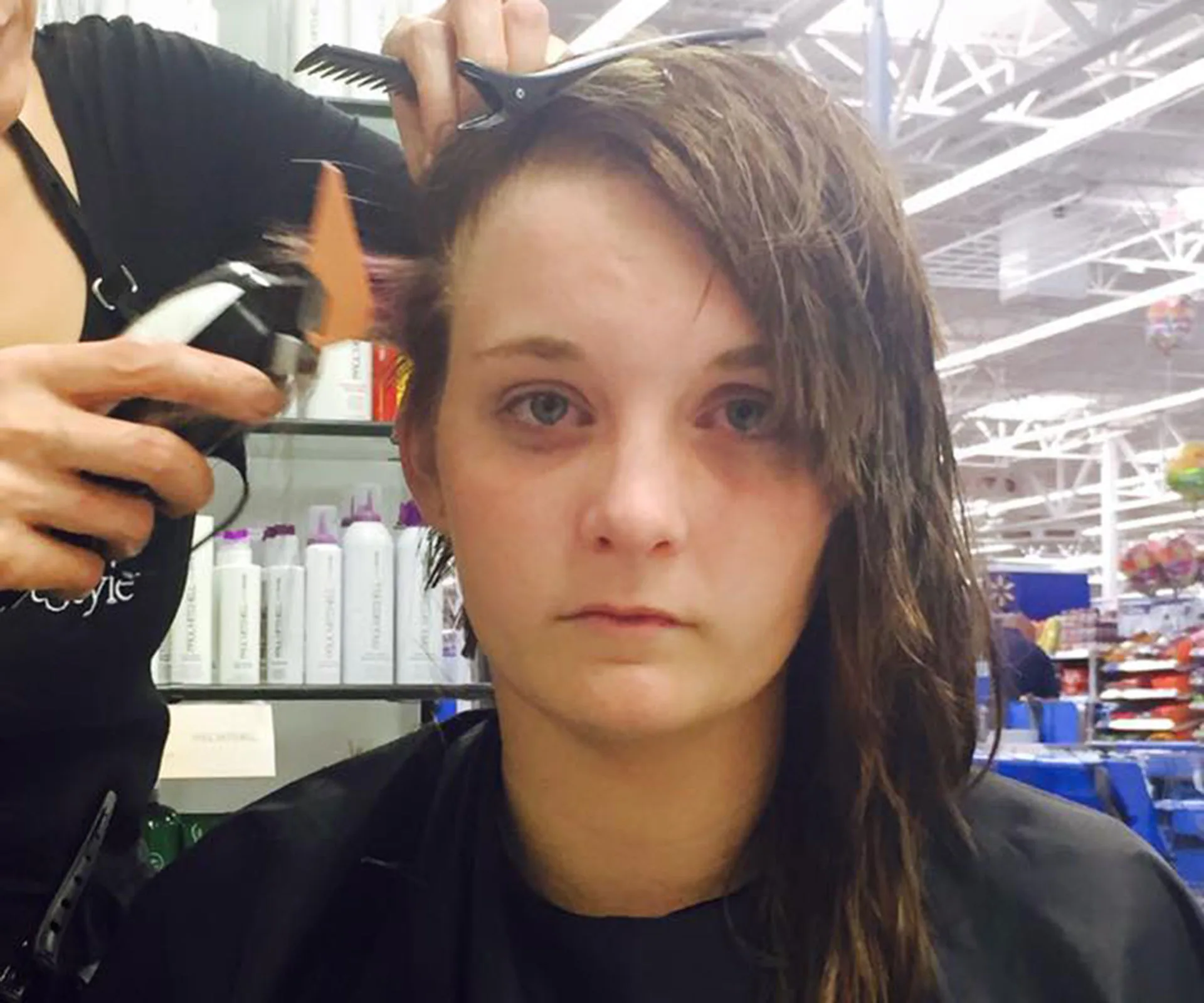 A girl with long hair looks solemn as a hairstylist starts to shave her head at a salon.