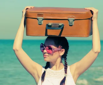 Woman in sunglasses holds a suitcase on her head at a beach.