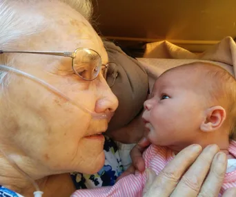 Elderly woman with glasses and oxygen tube gazes closely at newborn baby, creating a tender, heartwarming moment.