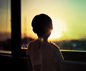 Child silhouetted against a sunset seen through a window.