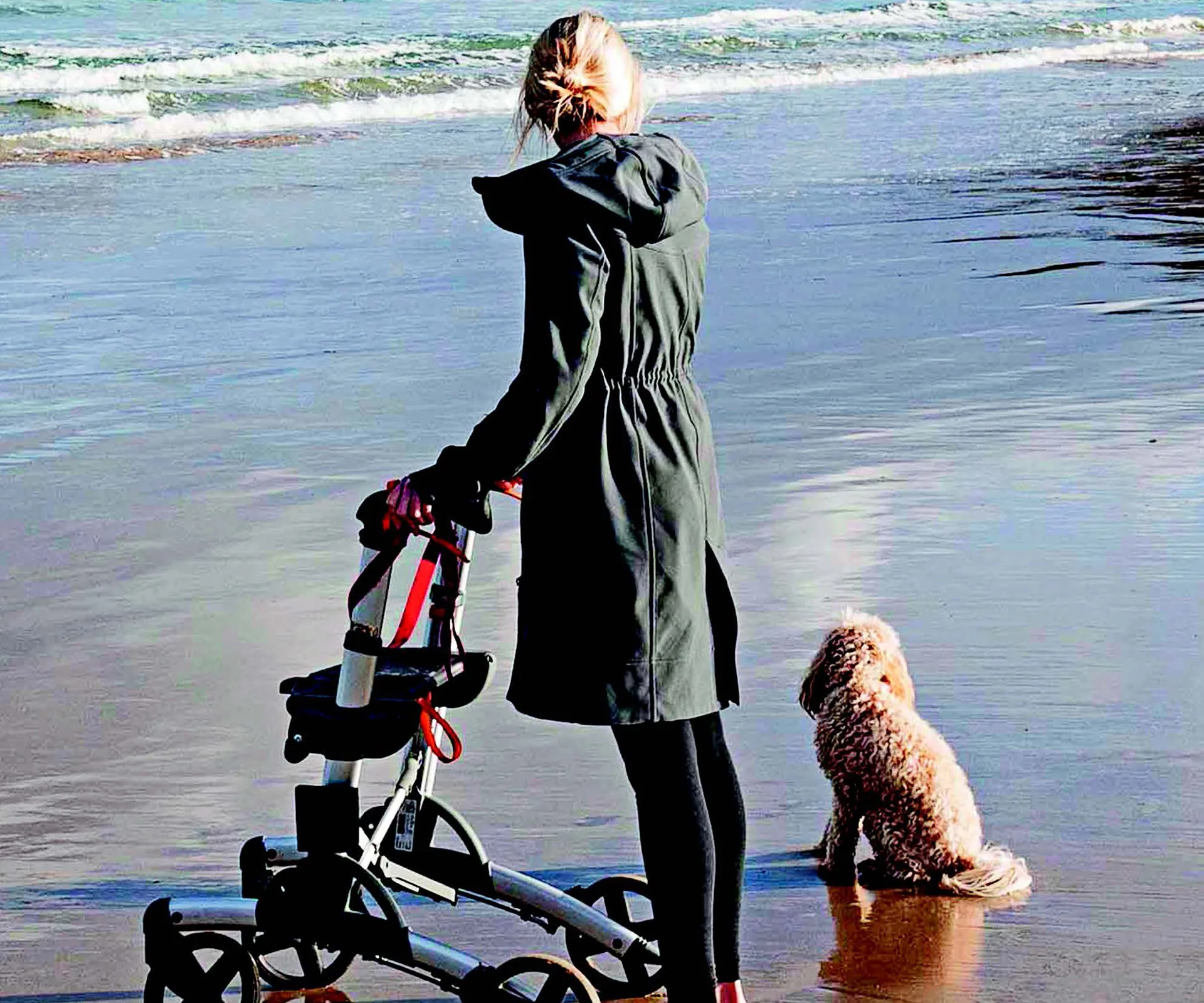 Woman with walker and dog on beach, facing the sea, wearing a green coat.