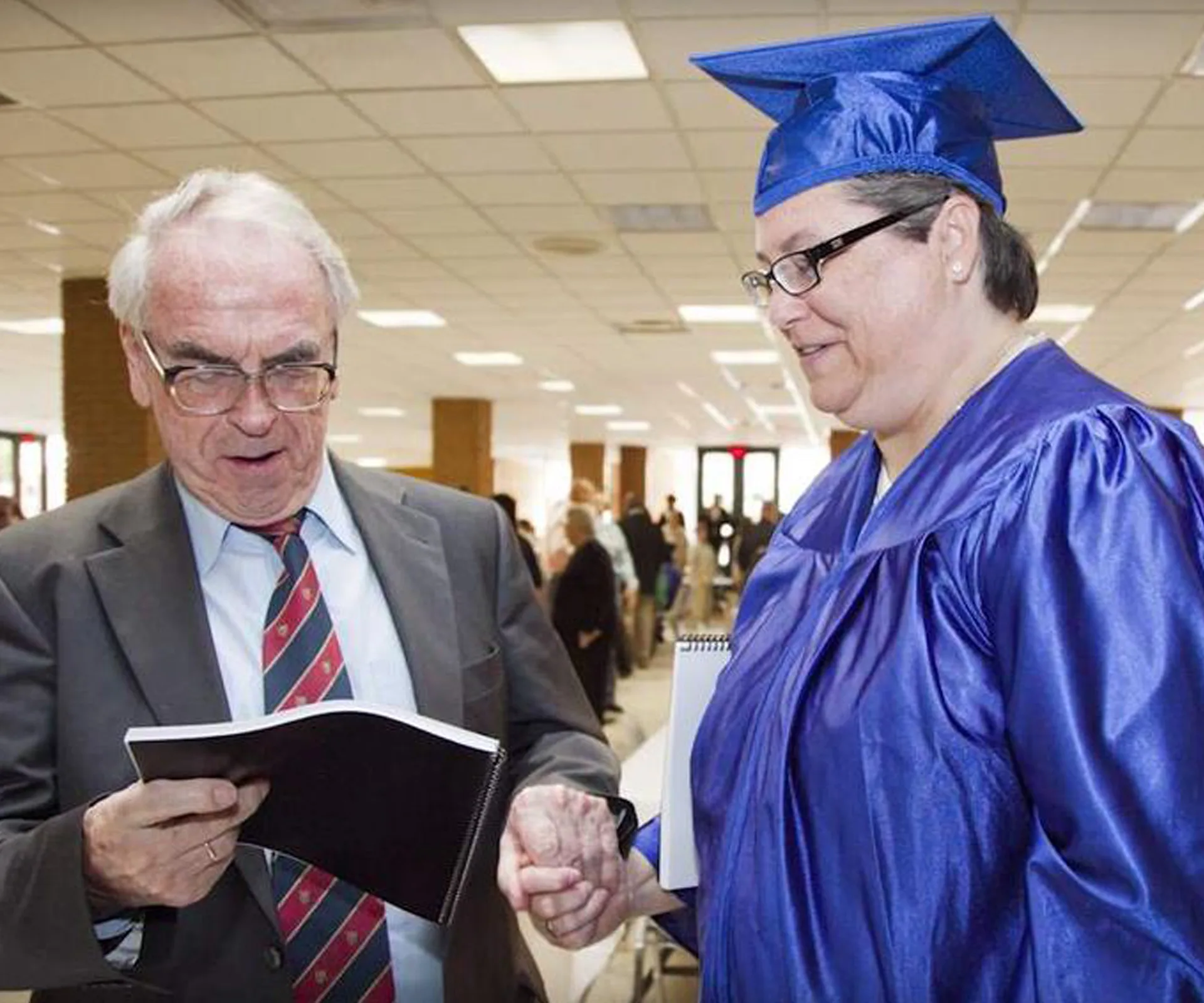 Man in suit and woman in blue graduation gown shake hands indoors.