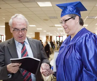Man in suit and woman in blue graduation gown shake hands indoors.