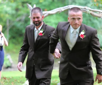 Father and stepfather in suits walk hand in hand at an outdoor wedding.