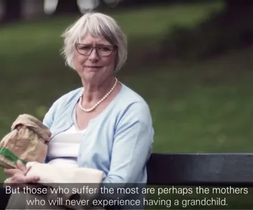 Elderly woman with gray hair and glasses sits on a park bench, holding a bag, wearing a blue cardigan and pearl necklace.