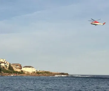Helicopter flying over a popular Sydney coastline, with buildings and people visible near the shore.
