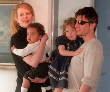 A couple holding two children in a warmly lit room; one adult wears sunglasses.