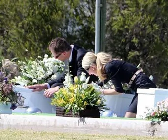 A man and woman in formal attire lean over a child-sized white coffin surrounded by flowers at a cemetery.