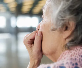 Elderly woman in a thoughtful pose, looking away with her hand near her mouth, in a softly lit room.