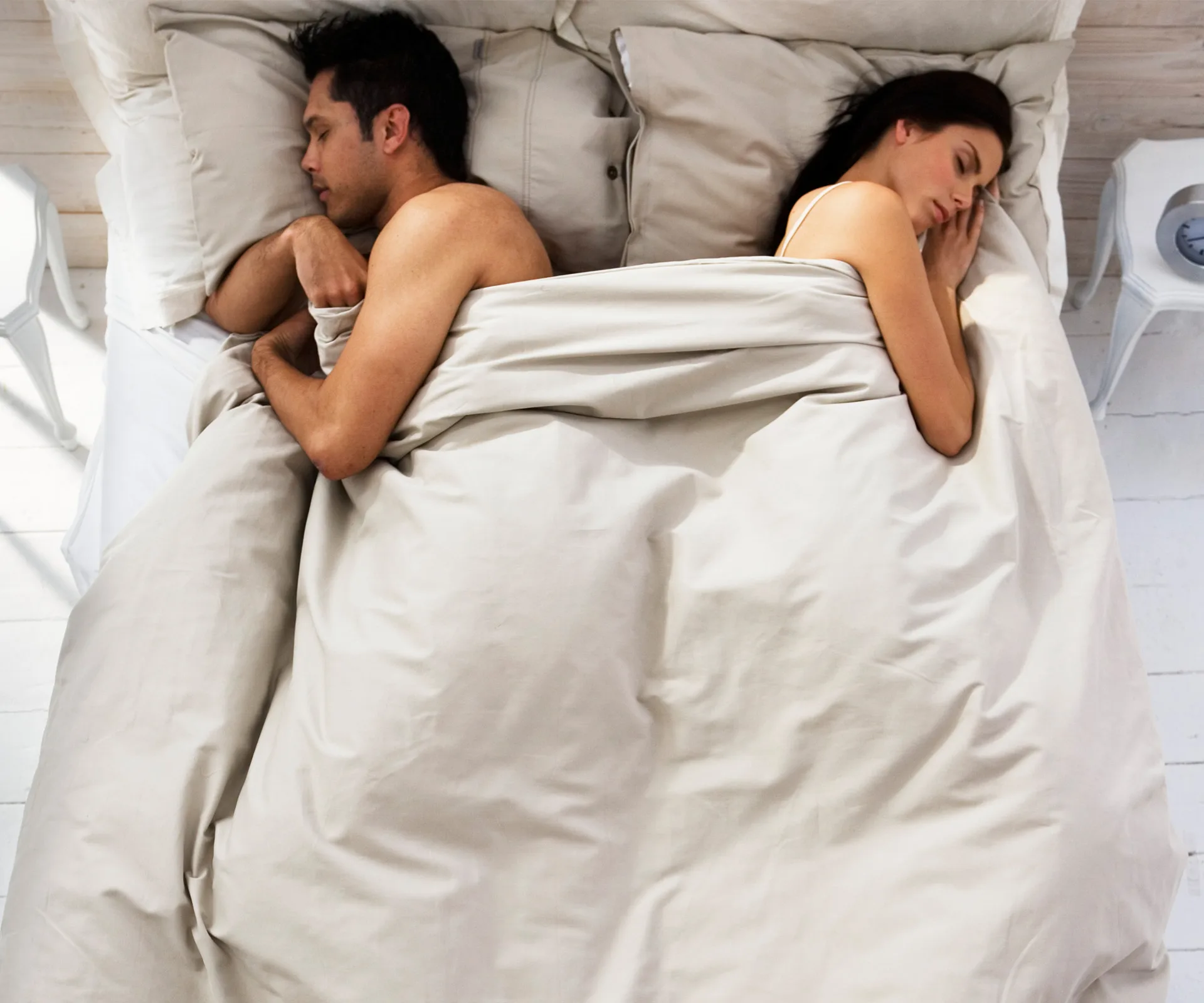 A couple lying back-to-back in bed, both asleep under beige sheets with neutral expressions, indicating possible distance.