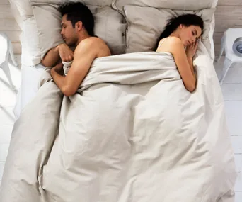 A couple lying back-to-back in bed, both asleep under beige sheets with neutral expressions, indicating possible distance.