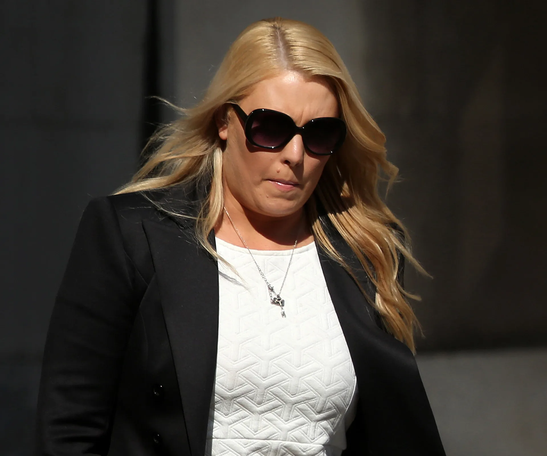 Woman with long blonde hair and sunglasses, wearing a white top and black coat, walking outdoors.