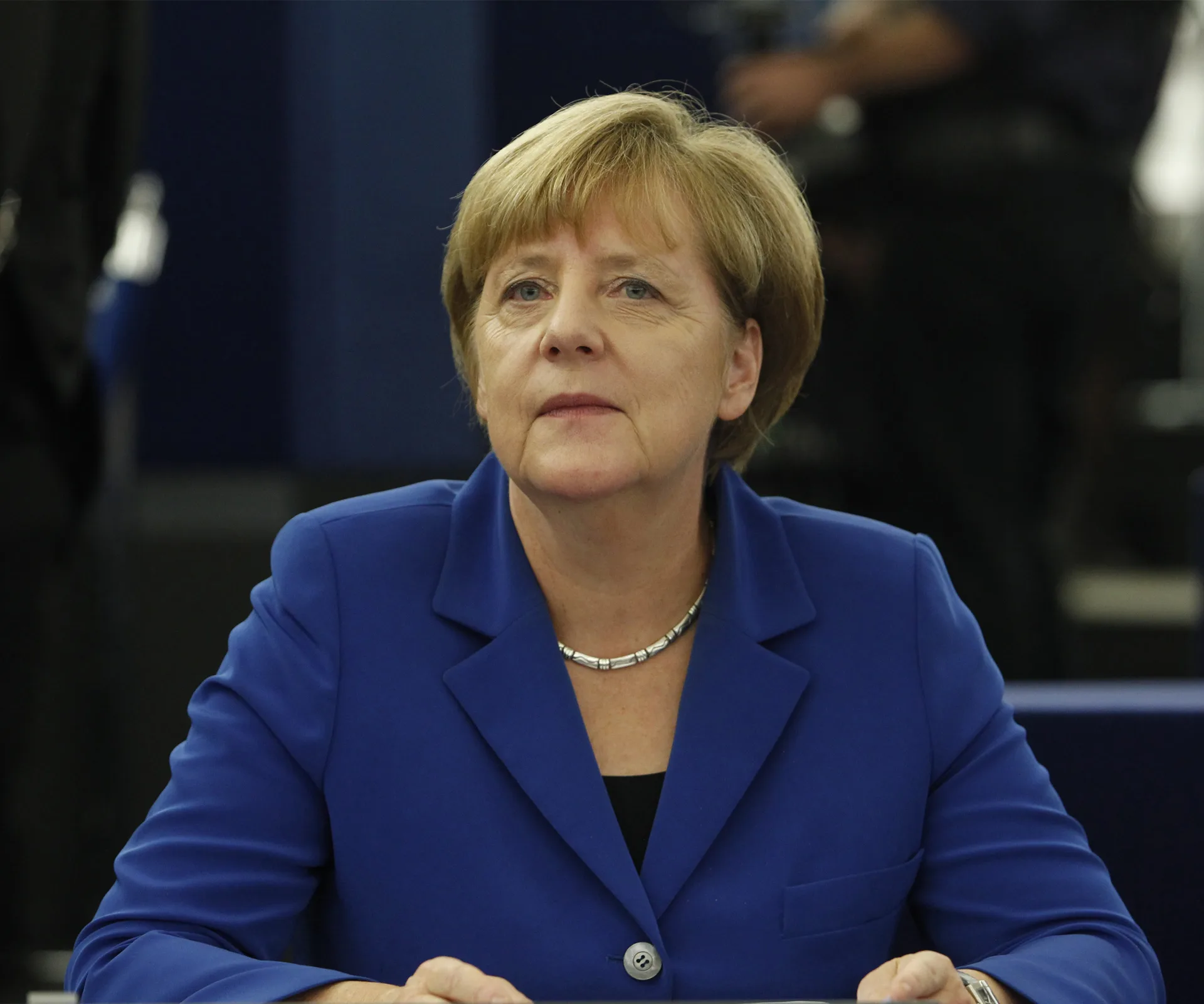 A person in a blue suit looks thoughtful, sitting at a table during an event.